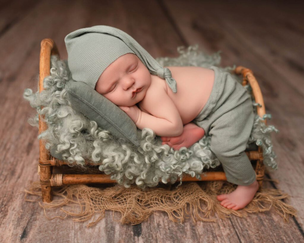two week old Baby boy wears sage green pants and matching sleepy cap while laying  on a bamboo newborn bed. 