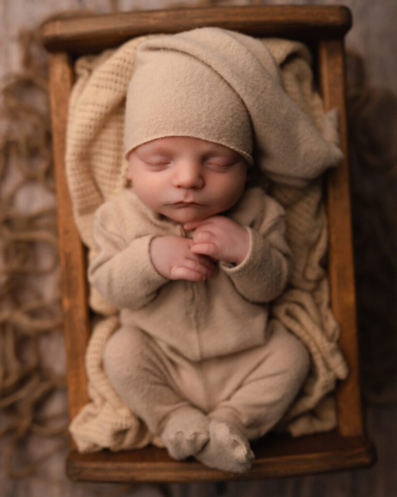 two week old baby boy wearing matching tan onesie and sleepy cap hat in a wooden bed on hardwood floors. 