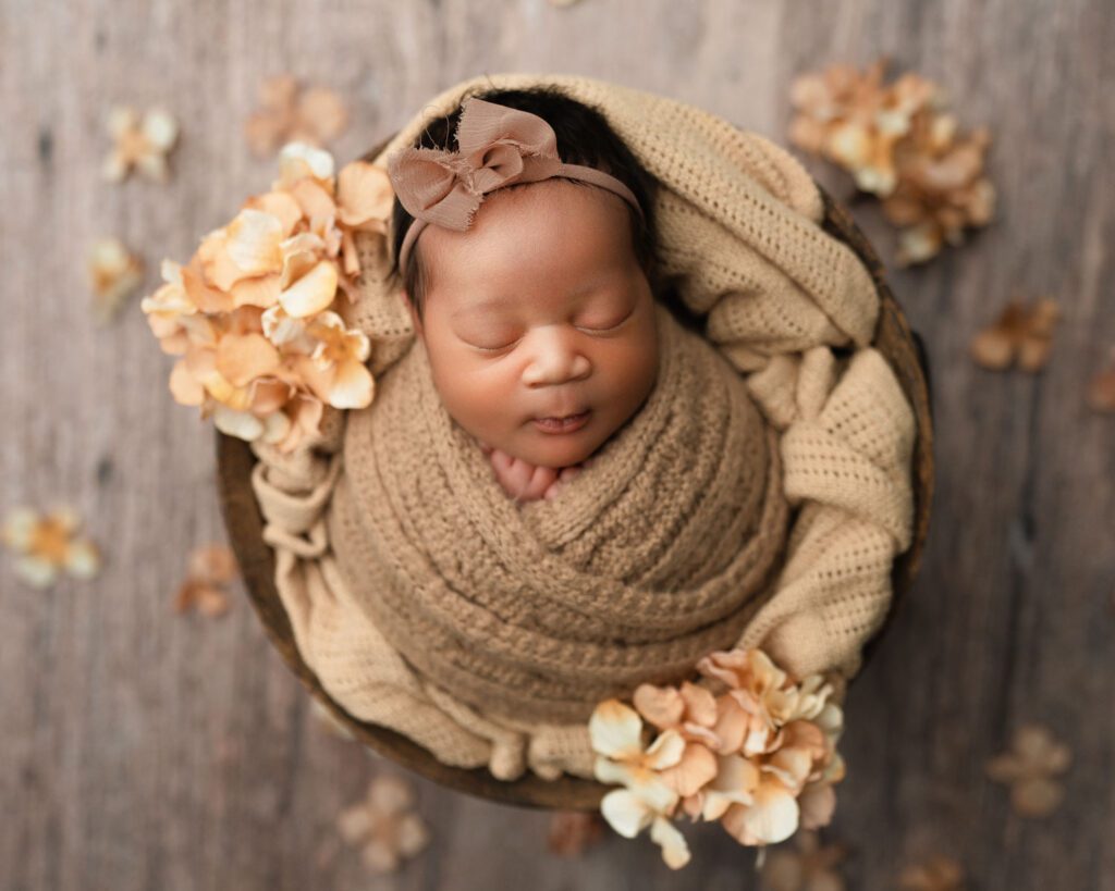 Brown blanket swaddles a newborn baby girl wearing a brown bow headband laying in a wooden bucket with tan flowers. 