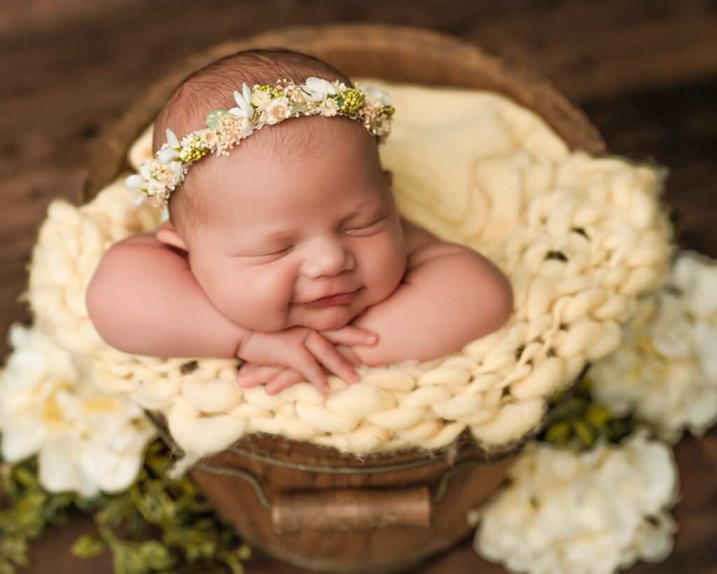 Smiling newborn baby girl wearing a yellow and green floral headband sits in a wooden bucket with yellow flowers around. 
