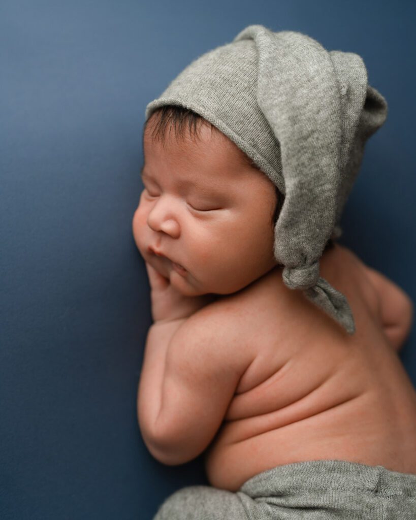 Sleeping newborn baby boy lays on his stomach on a blue backdrop and wears grey pants and a matching sleepy cap hat. 