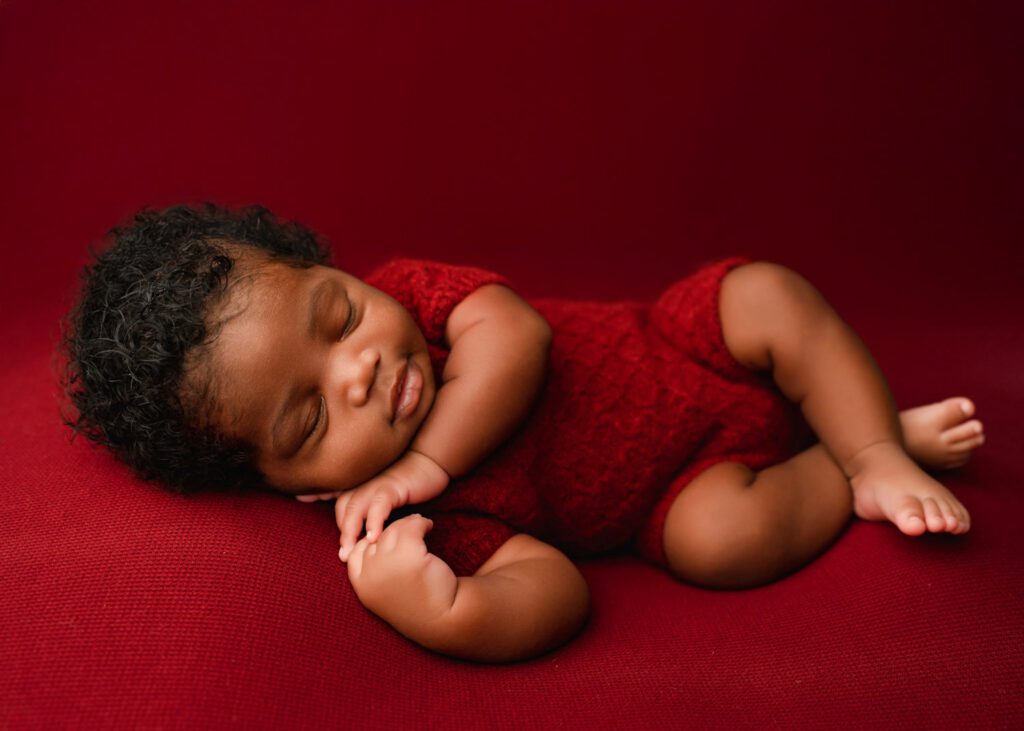Baby boy lays on his side with hands by his face wearing a red textured outfit laying on a red blanket background. 