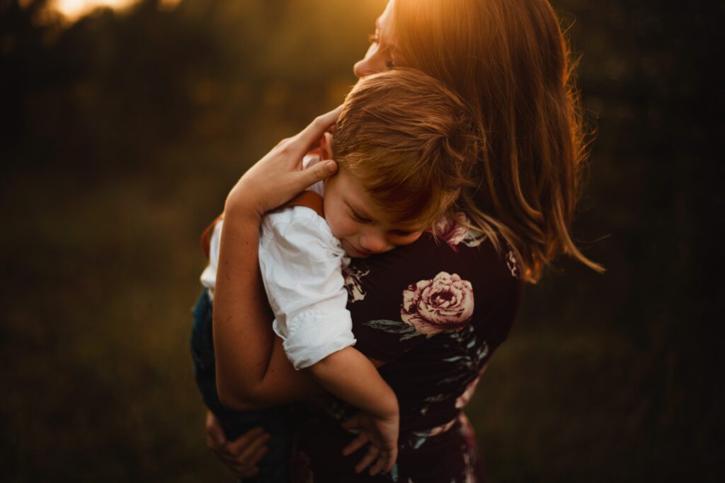 Young boy wears a white shirt and brown suspenders is upset and crying while his mother holds him with her hand on his head. 