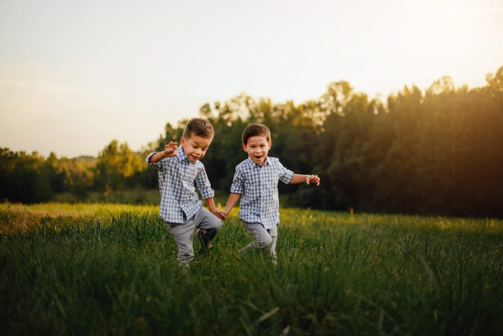 Twin boys wearing matching blue and white plaid shirts act silly and pretend to be dinosaurs while walking through grass. 
