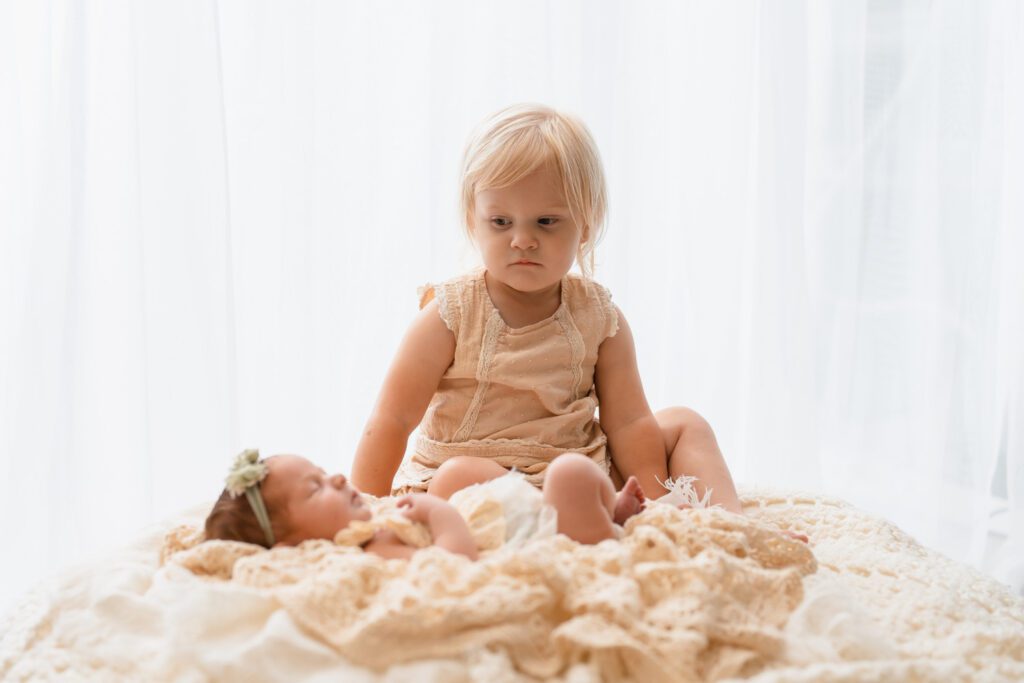 Big sister with an unsure look on her face looks down at her newborn baby sister sleeping while laying on white blankets. 