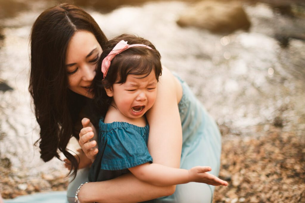 Little girl wearing a blue jean dress and pink bow in her hair is crying with adorable hand gesture as her mom comforts her.