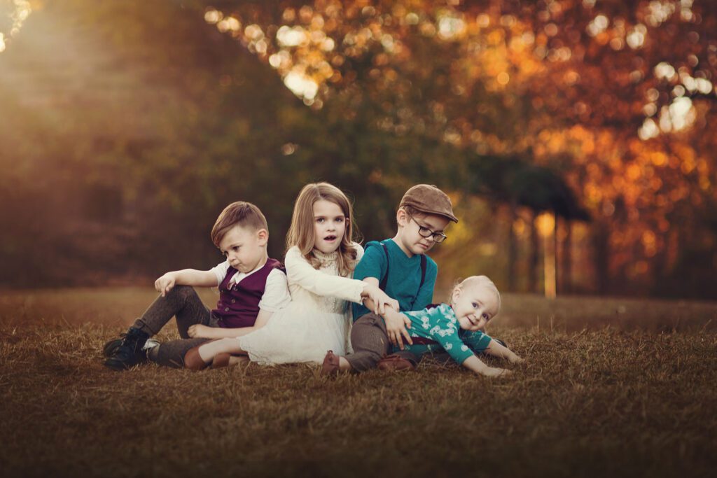Four siblings sit in the grass as the youngest baby tries to escape while older sister has a shocked look on her face. 